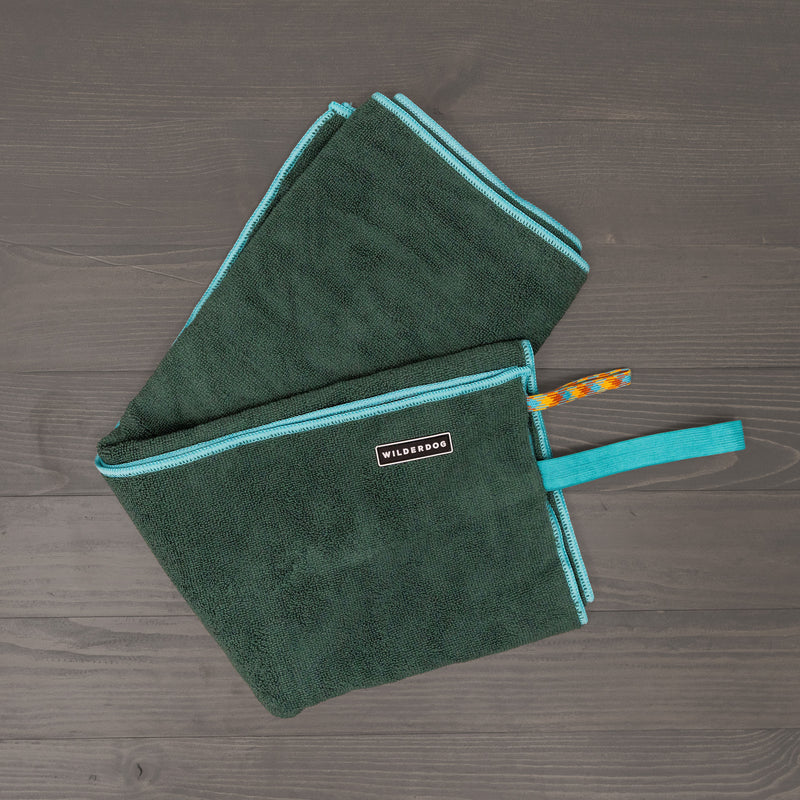 The Quick Dry Towel, made of dark green polyester with light blue trim and zipper, features a "WILDS&DOGS" label and colorful hanging loop, shown on a dark wooden surface.